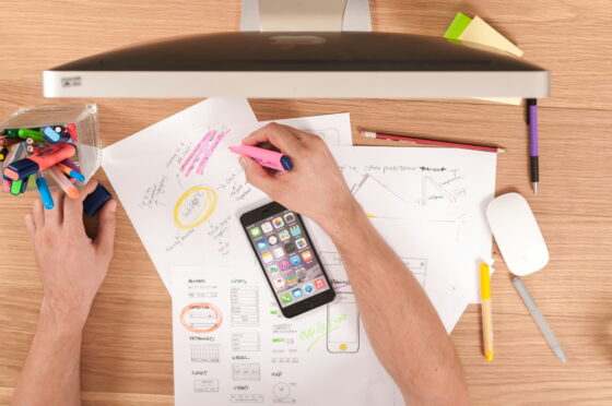 Person using a smartphone and highlighters while reviewing sketches and plans on paper in a home office, symbolizing strategic planning for Amazon affiliate marketing success in 2026.