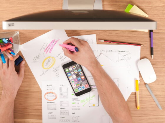 Person using a smartphone and highlighters while reviewing sketches and plans on paper in a home office, symbolizing strategic planning for Amazon affiliate marketing success in 2026.